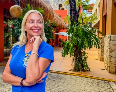 A smiling woman with blonde hair, wearing a blue t-shirt, stands outdoors in a sunny, tropical courtyard with orange buildings and lush green plants. She is looking thoughtfully away from the camera, her hand resting under her chin, exuding a clear sense of pride and confidence in her new smile.
