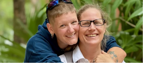 A medium close-up portrait of two smiling women embracing outdoors in a tropical, leafy setting.
