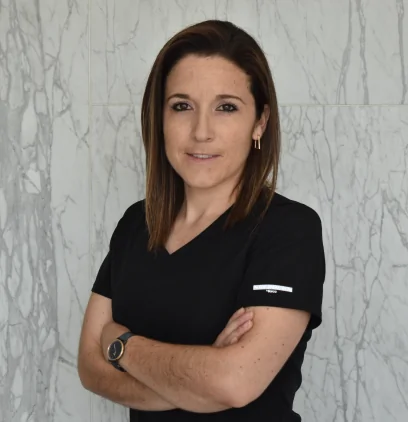 Portrait of Dr. Alejandra Paz, dental specialist, wearing black scrubs and standing with arms crossed against a marble wall.