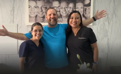 Enthusiastic male patient with arms outstretched in celebration, posing with Dr. Martha Tziu and a dental assistant inside the modern Smile Makeover clinic.