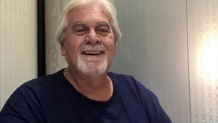 smiling older man with white hair and a white goatee wearing a dark blue t-shirt. He is showing his teeth in a warm smile.