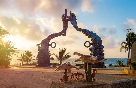 Portal Maya sculpture in Playa del Carmen, featuring two stylized figures forming an arch, with sea turtles and other marine life sculptures in the foreground against a coastal sunset.